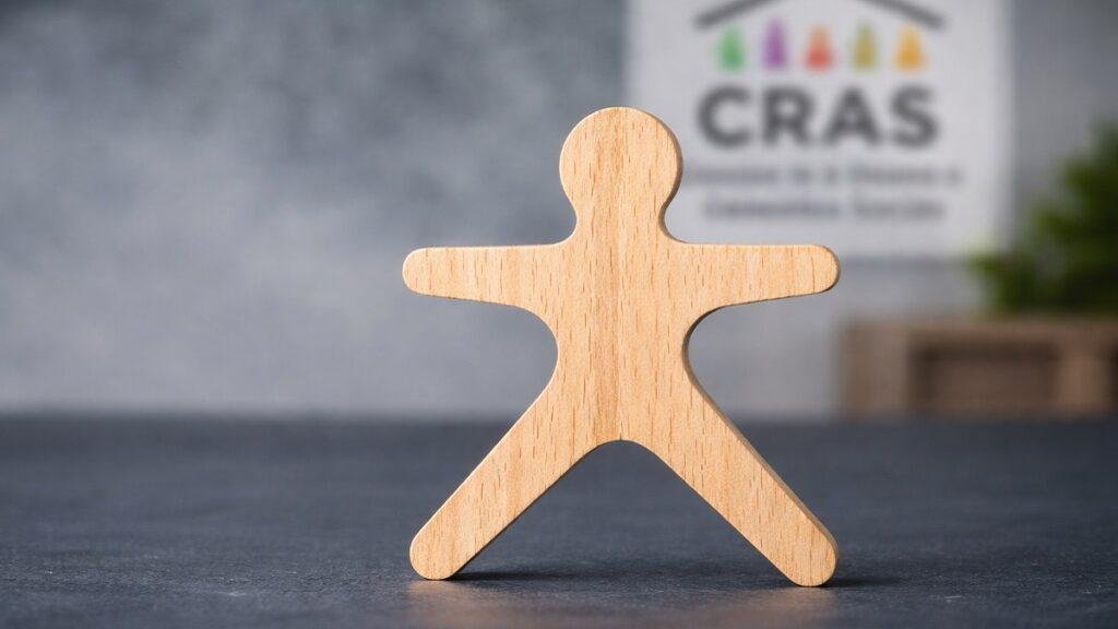 Figura de madeira representando pessoa sozinha com unidade do CRAS desfocada ao fundo, simbolizando família unipessoal e acesso a benefícios sociais.