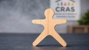 Figura de madeira representando pessoa sozinha com unidade do CRAS desfocada ao fundo, simbolizando família unipessoal e acesso a benefícios sociais.